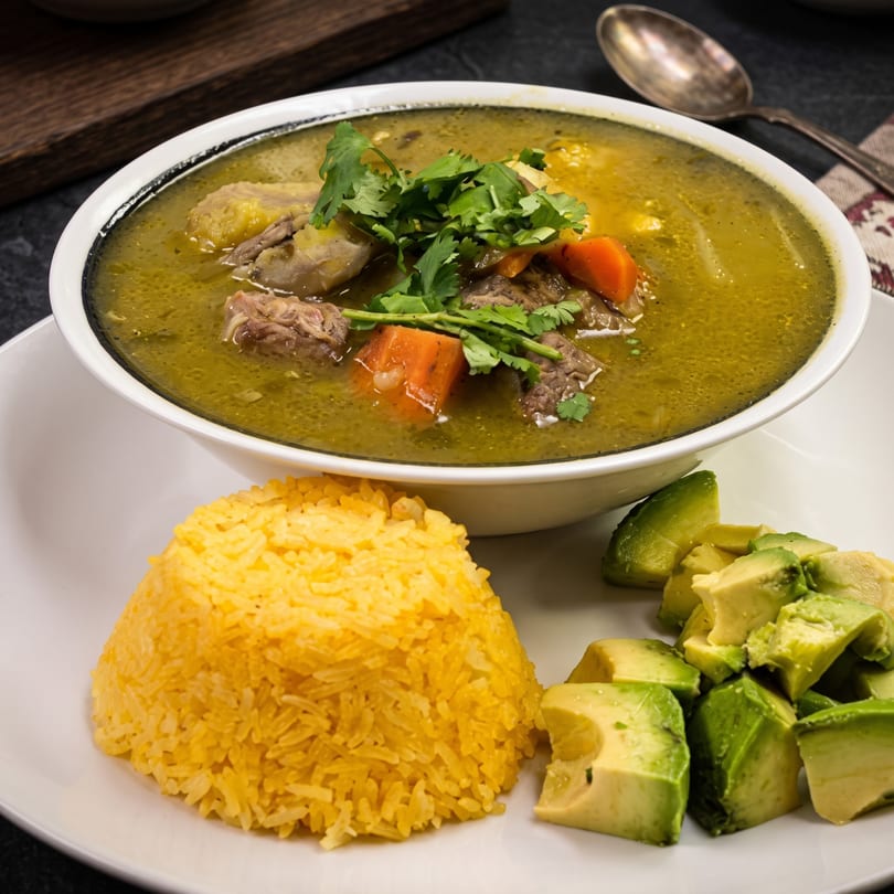 Hearty green soup with meat, carrots, and cilantro, served with yellow rice and diced avocado.