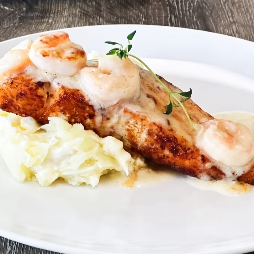 Pan-seared salmon topped with shrimp and creamy sauce, served with mashed potatoes and a sprig of thyme.