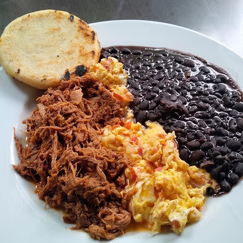 A white plate with shredded beef, scrambled eggs, black beans, and an arepa.