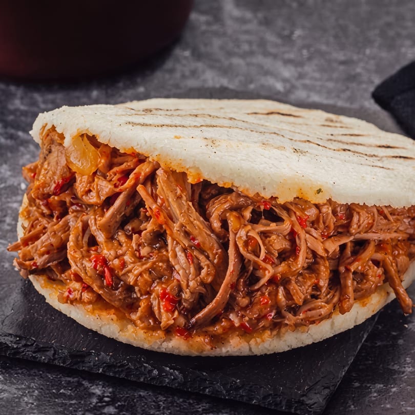 Close-up of a Venezuelan arepa filled with shredded beef and red peppers on a dark surface.