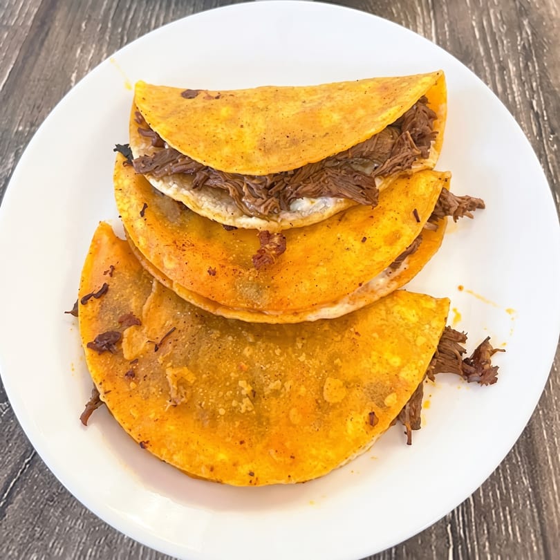 Three birria tacos with shredded beef and melted cheese on a white plate.