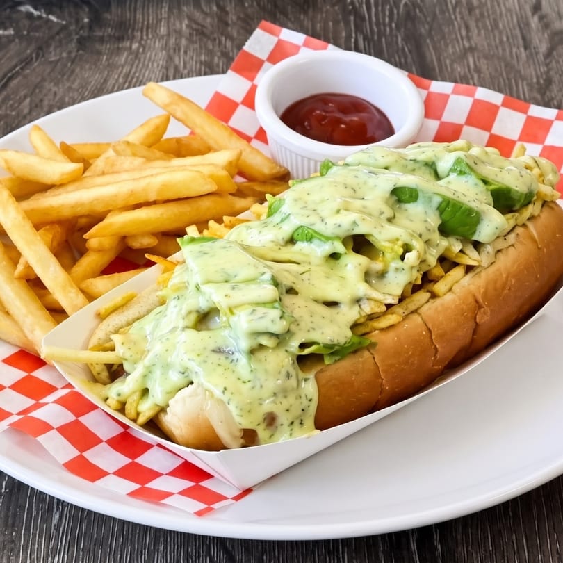 Hot dog with green sauce and avocado, served with fries and ketchup on a checkered paper.