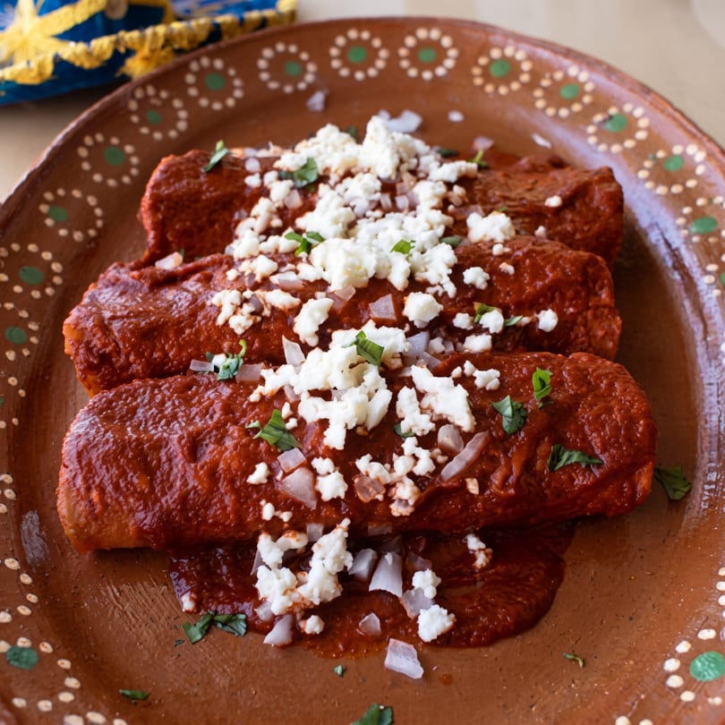Three red enchiladas topped with crumbled white cheese, diced onions, and cilantro on a brown plate.