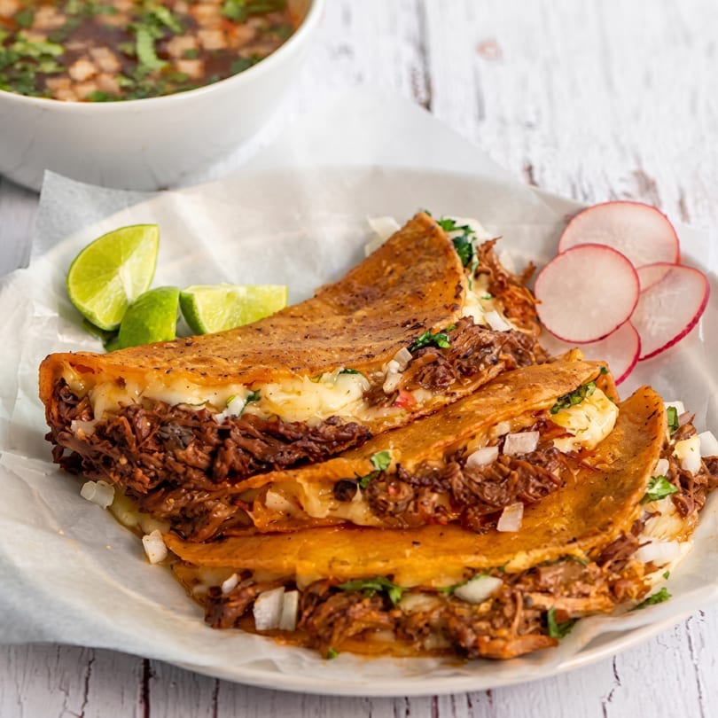 Three birria tacos with melted cheese, onions, and cilantro, served with lime wedges and sliced radishes.