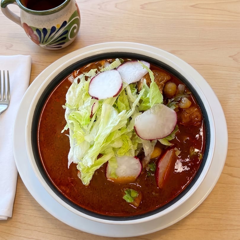 A bowl of pozole rojo with shredded lettuce and radish slices, next to a decorative mug.