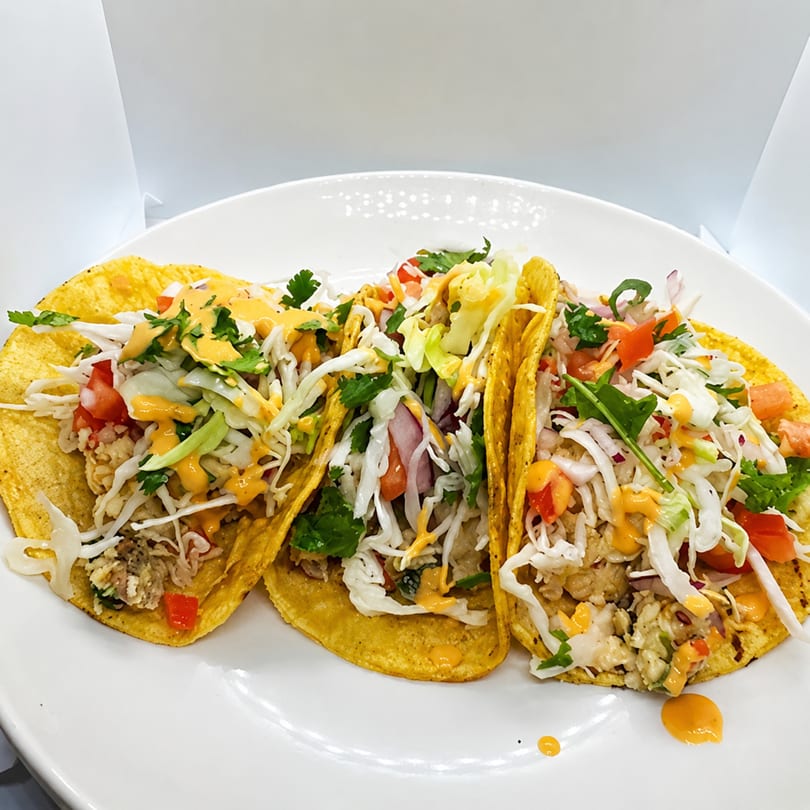 Three vibrant tacos filled with meat, shredded cabbage, diced tomatoes, and a creamy orange sauce.
